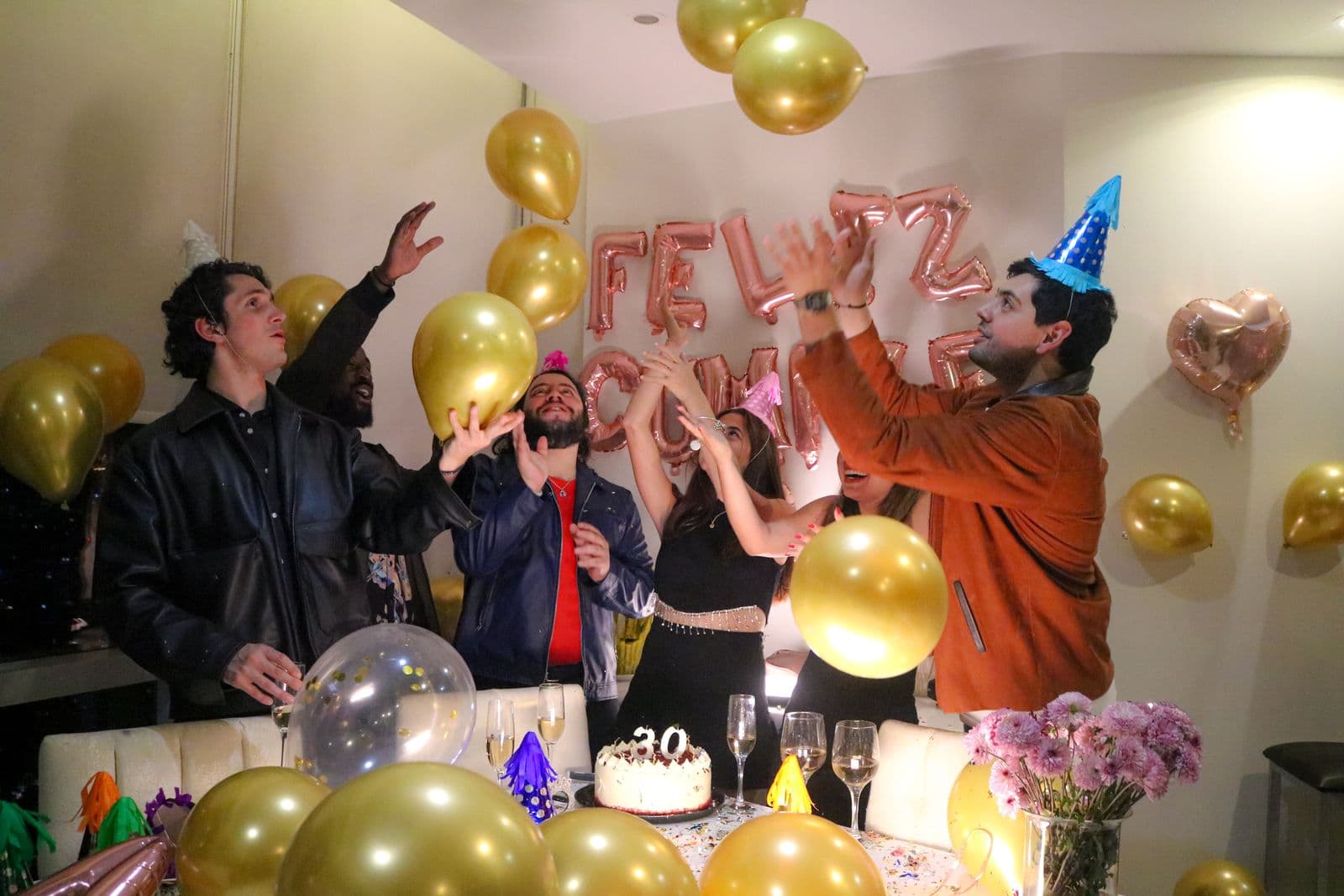 Friends tossing gold balloons at a 30th birthday party with a cake on the table