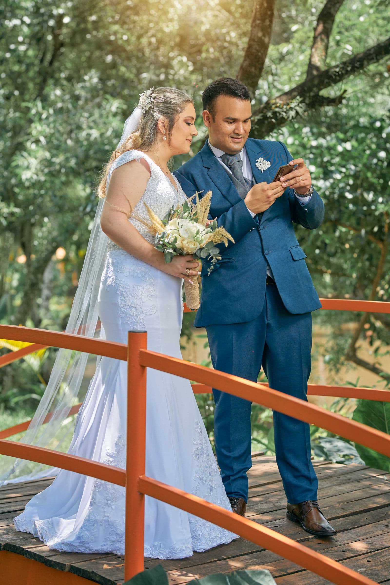 A bride and groom looking at a phone together on a wooden bridge
