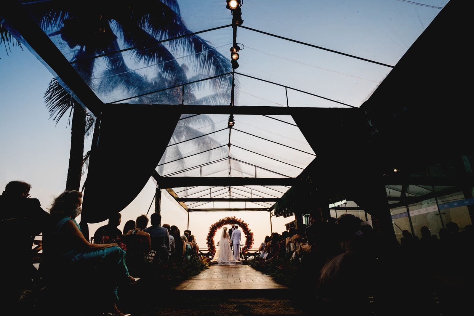 Guests watching an evening wedding ceremony under a glass tent at dusk