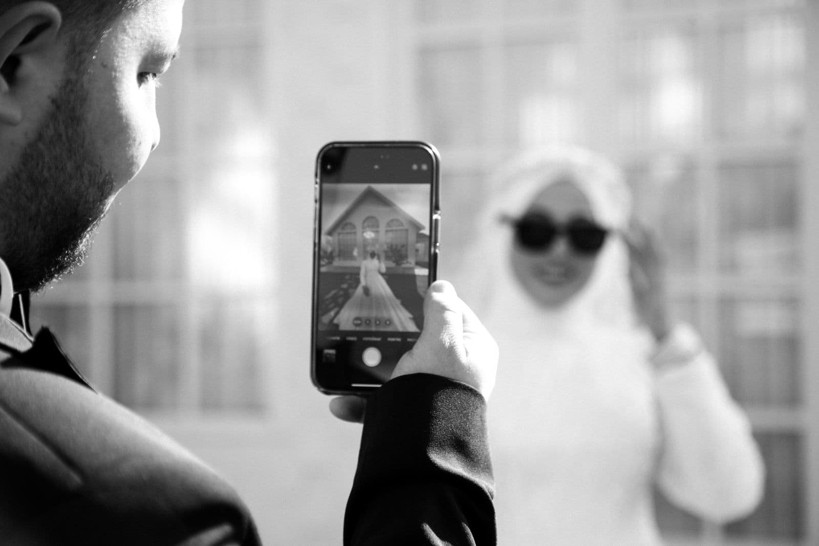 A groom photographing his bride on a phone in black and white