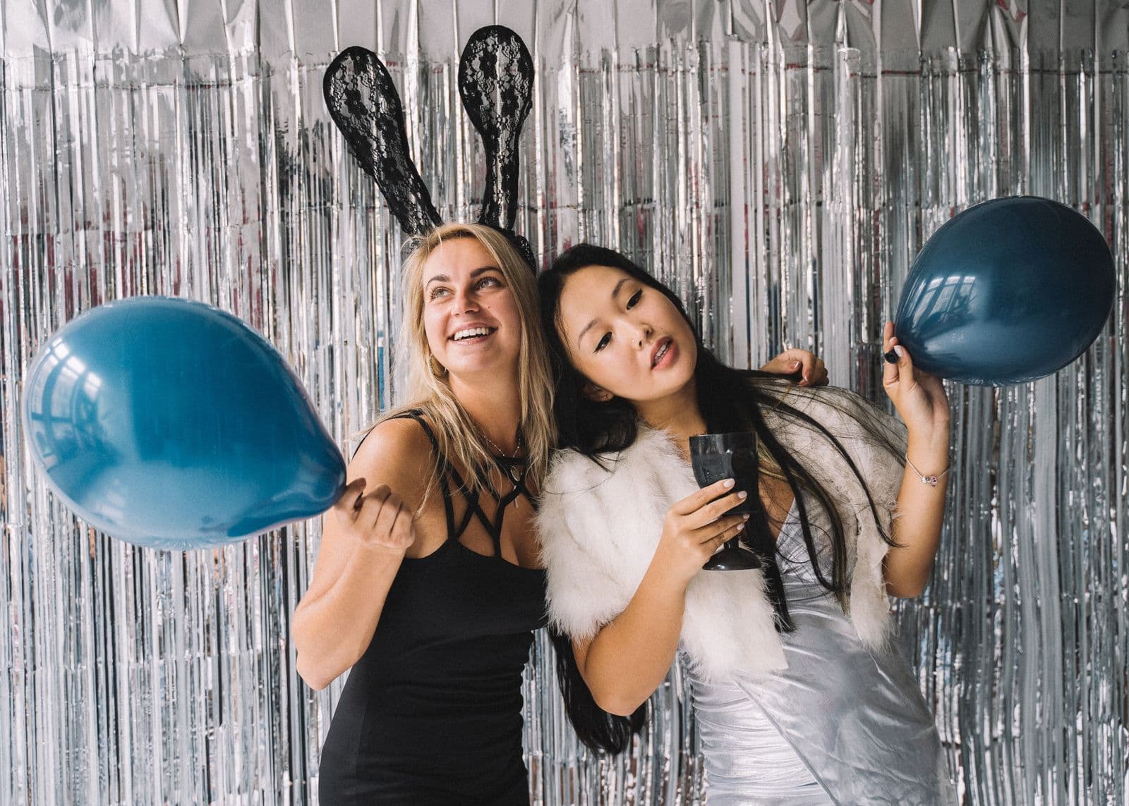 Two friends posing with balloons in front of a shiny party photo-booth backdrop